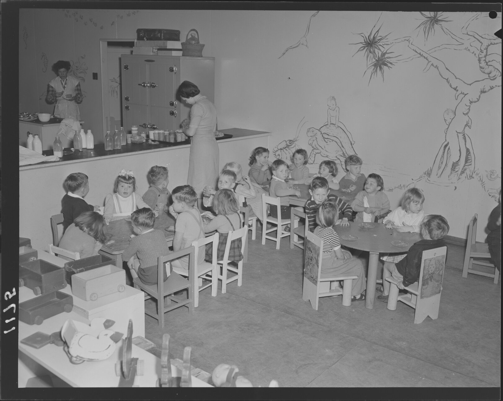 Children at tables eating, teachers at kitchen at Kindercraft Civic Nursery (childcare) City Hall - 1950