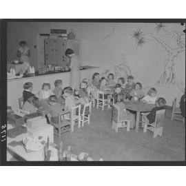 Children at tables eating, teachers at kitchen at Kindercraft Civic Nursery (childcare) City Hall - 1950